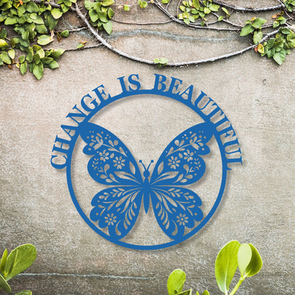 Blue butterfly-shaped metal sign with 'Change is Beautiful' text on a stone surface with greenery.
