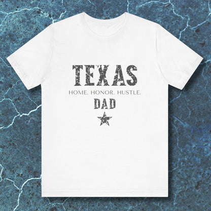 White t-shirt with 'TEXAS HOME HONOR HUSTLE DAD' text on a textured blue background