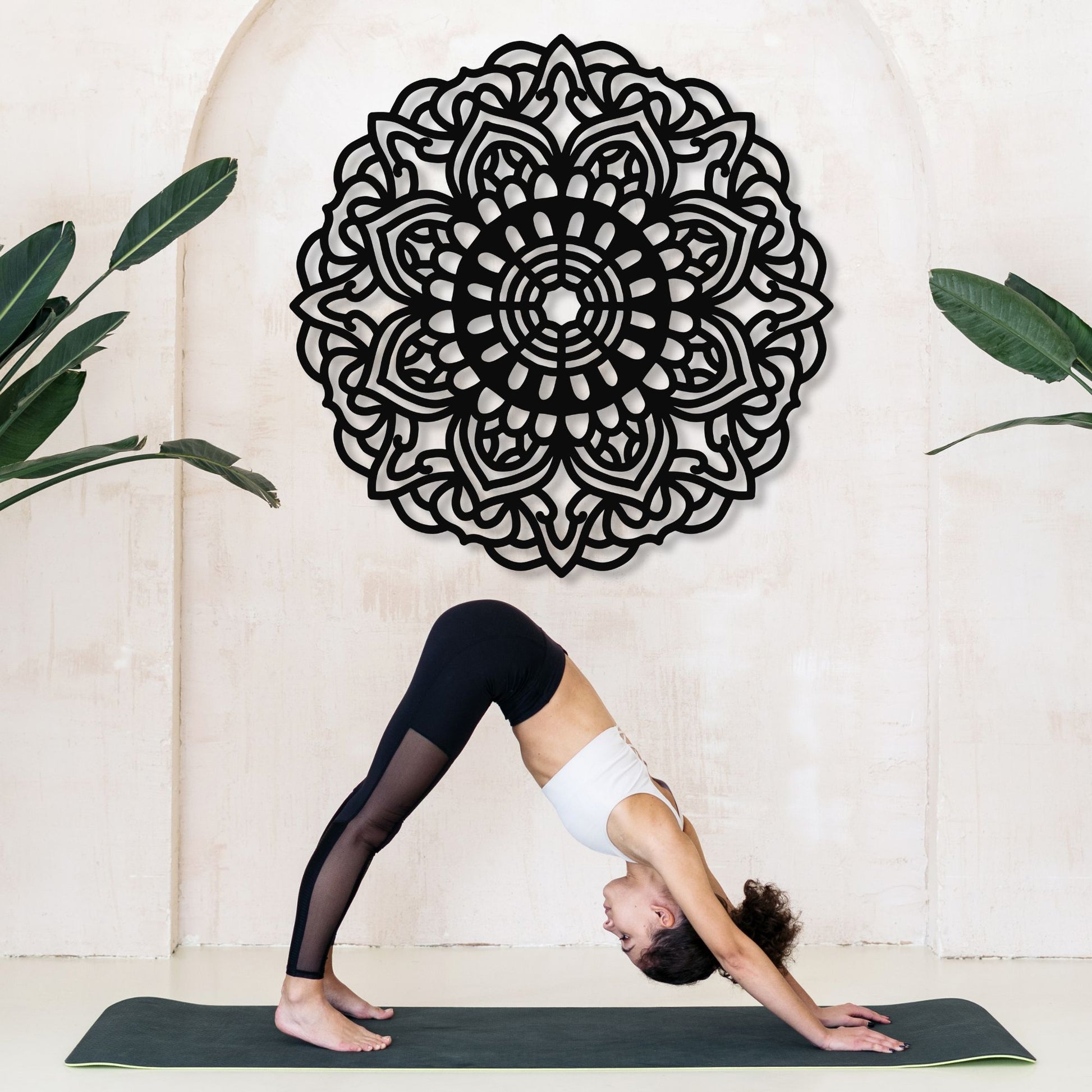 Person practicing yoga in front of a decorative mandala wall art with plants on either side.